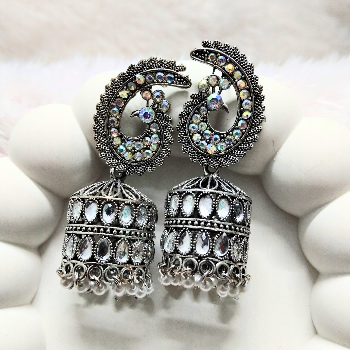 Oxidized jhumka earrings with peacock head design and sparkling stones, placed on a soft white cushion