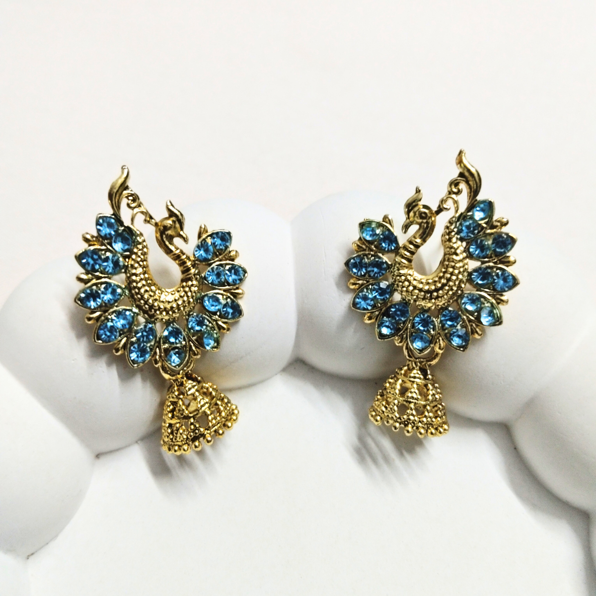 Gold peacock earrings with colored stones and jhumka danglers – shown in blue variant on a white background.