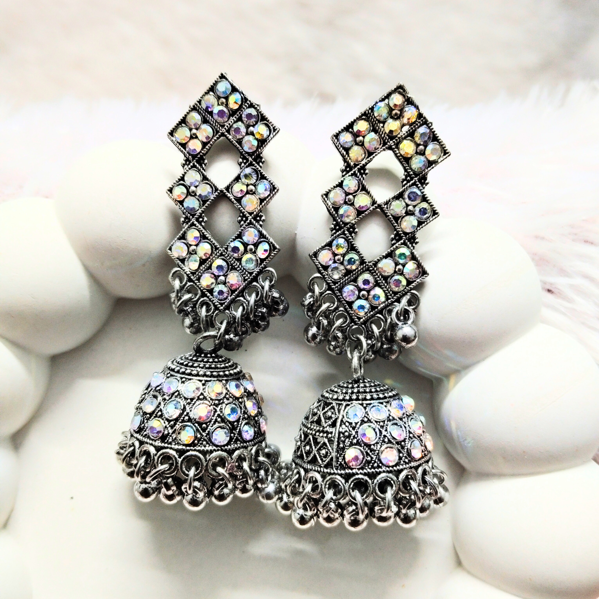 Oxidized silver jhumka earrings with diamond lattice top and stone embellishments placed on a white cushion background.