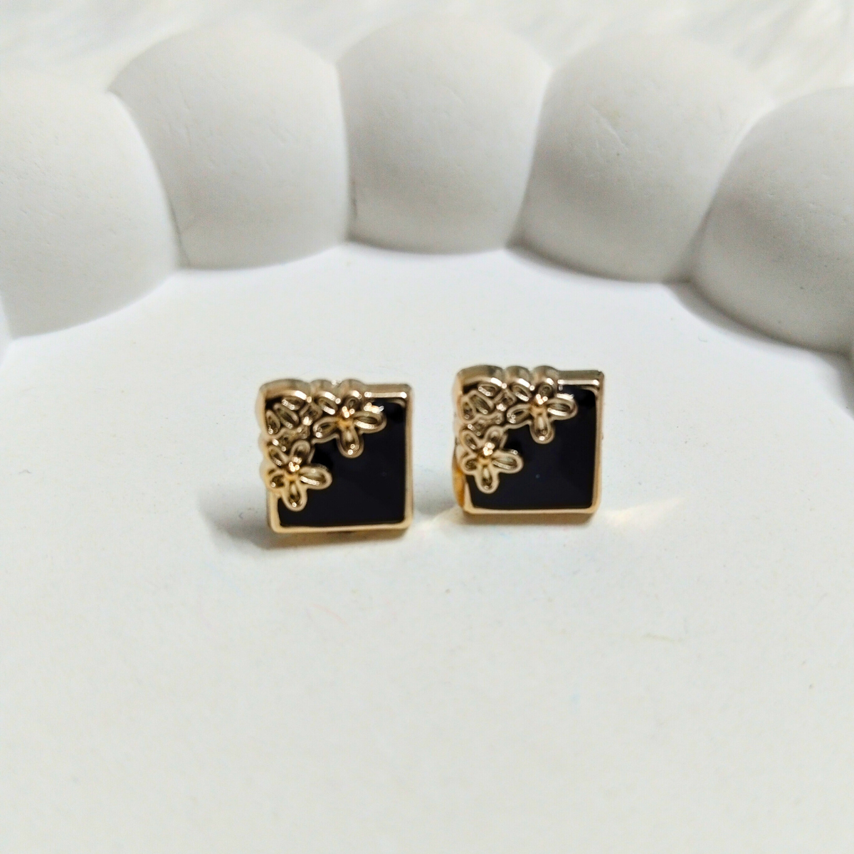 Gold square stud earrings with black enamel and floral detailing on a white surface