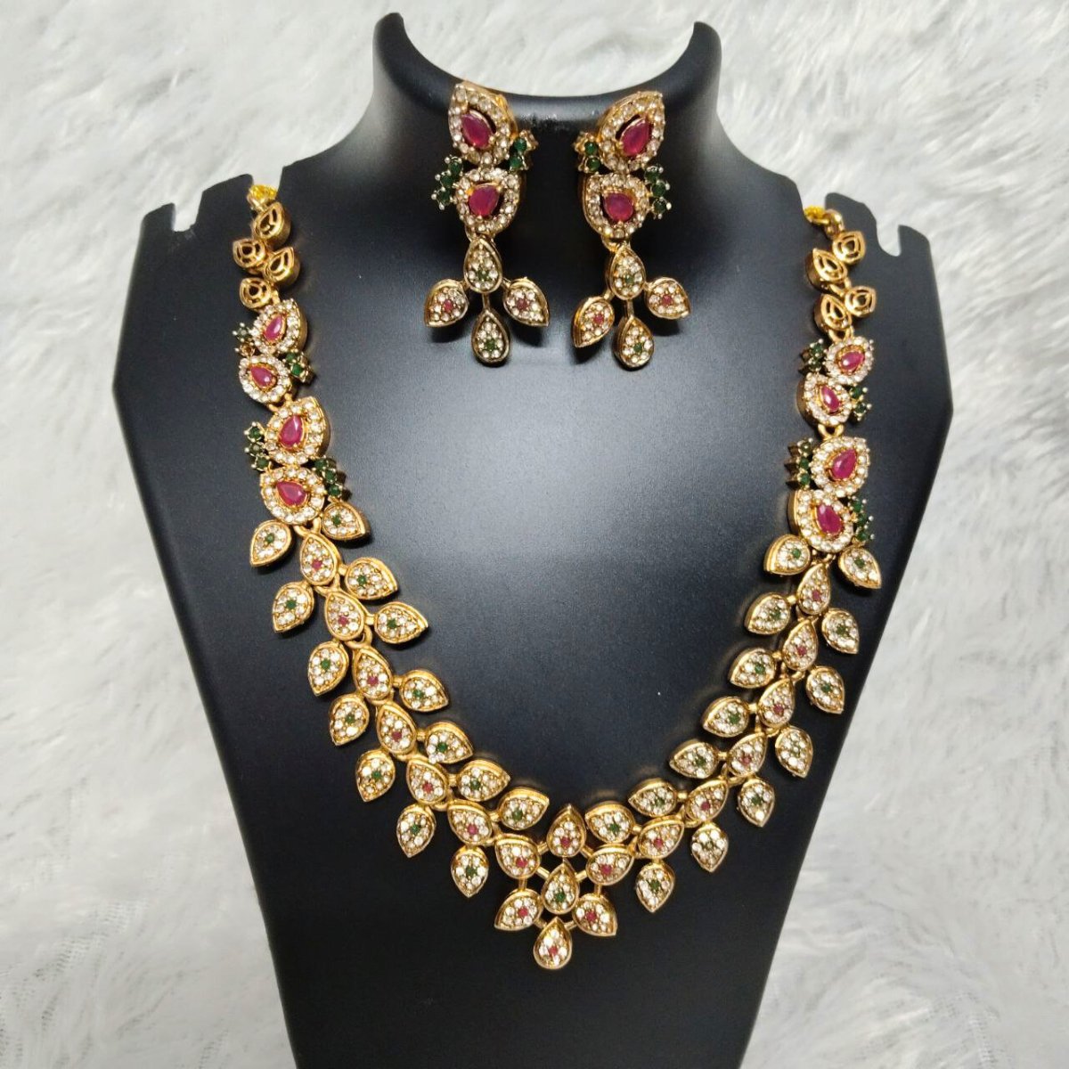 Gold plated necklace set with ruby red and green stones in a leaf design, paired with matching earrings, displayed on a black jewelry stand.